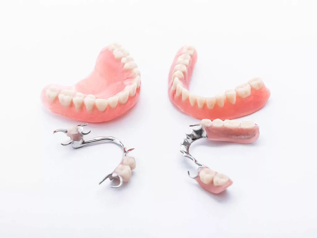 Two full sets of dentures and two partial dentures placed on a white background. The full dentures are pink with white artificial teeth, and the partial dentures have metal clasps with a few artificial teeth on a pink base.