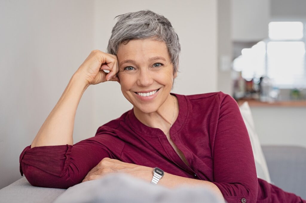 older woman smiling while sitting on a couch with her head on her hand