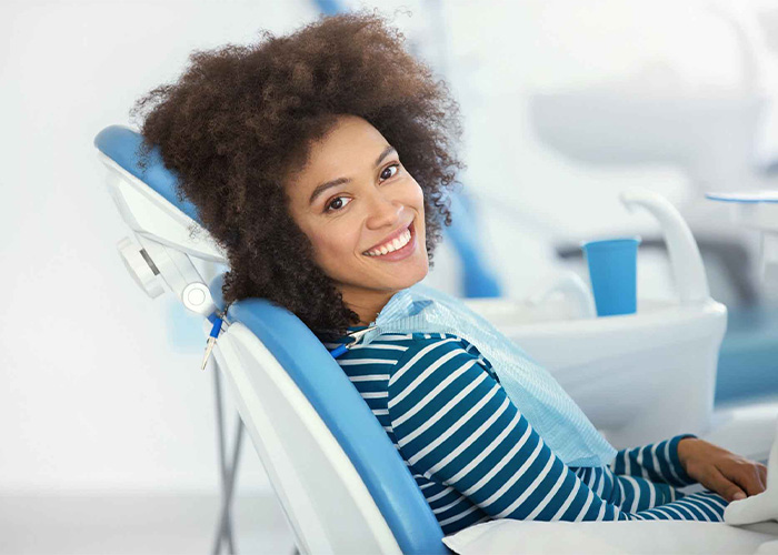 Patient sitting in a dental chair at a dentist's office, smiling, wearing a striped shirt and dental bib. Dental equipment and instruments are visible in the clinical setting.